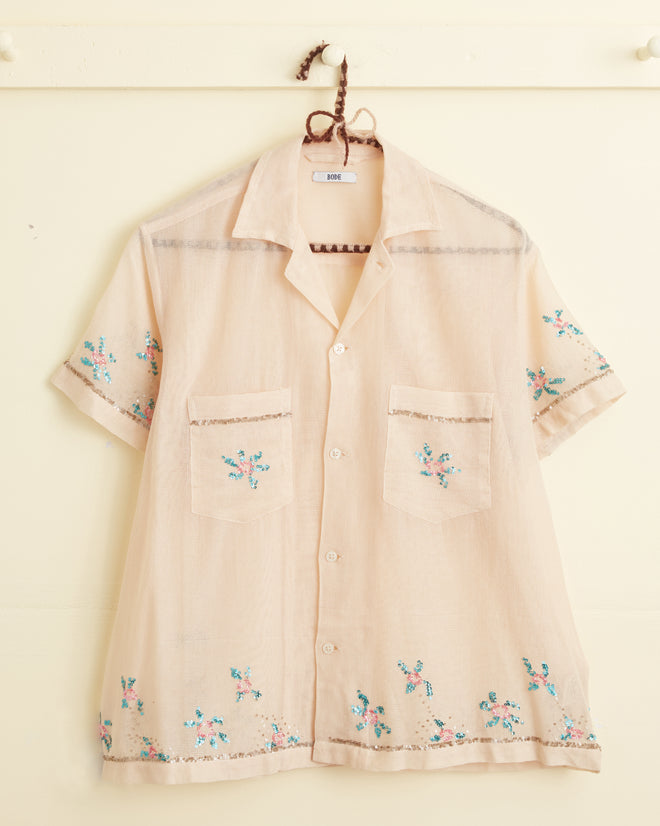 The Sequined Floral Net Short Sleeve Shirt is a light beige button-up with blue floral embroidery and an appliqué flower design. It features two front pockets and a collar, all showcased against a cream-colored background on a twisted string hanger.