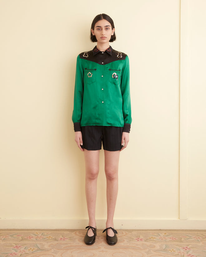 A person stands against a cream-colored wall, wearing the Sequin Rodeo Shirt in dark green with black shorts. The shirt features embroidery on the shoulders and pearl snap buttons. They have short hair and wear black shoes.