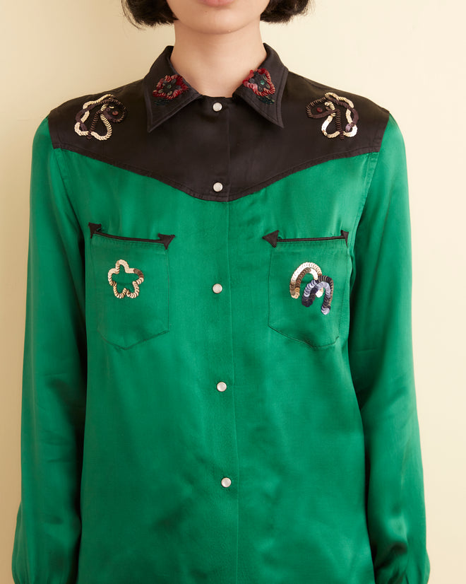 A person stands against a cream-colored wall, wearing the Sequin Rodeo Shirt in dark green with black shorts. The shirt features embroidery on the shoulders and pearl snap buttons. They have short hair and wear black shoes.