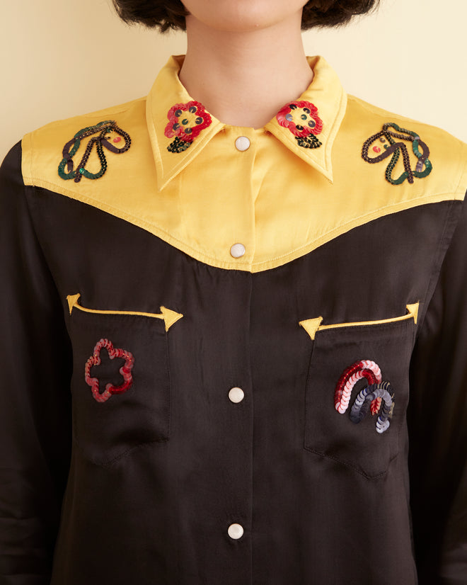 A person stands against a light-colored wall wearing a Sequin Rodeo Shirt in gold and black with pearl snap buttons, paired with black wide-legged pants. They are looking directly at the camera with a neutral expression.