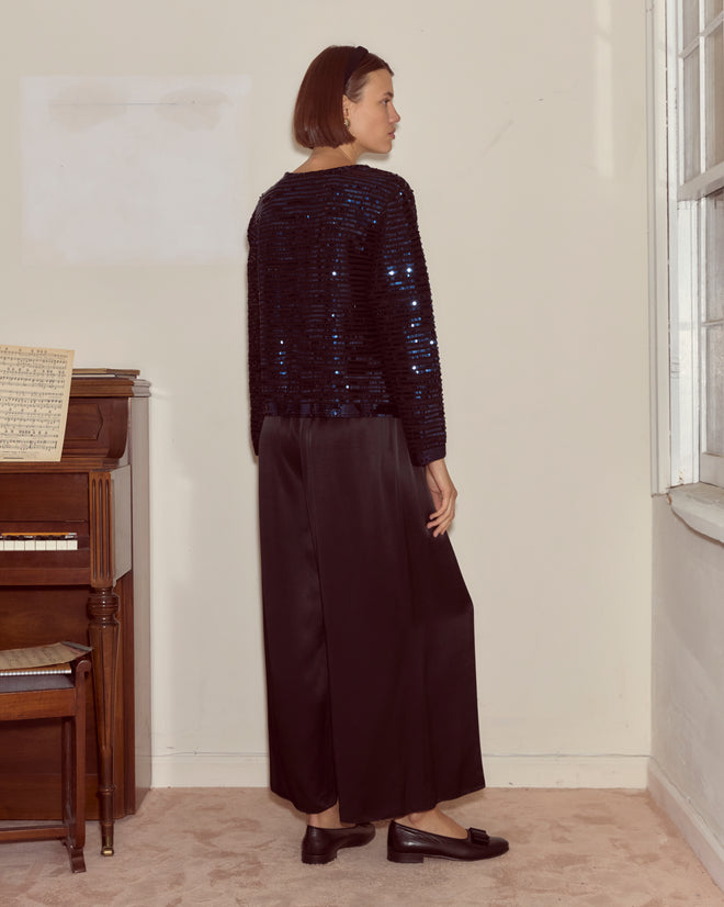 Woman in the Sequin Spotlight Swing Cardigan - Black/Blue stands by a piano, facing the wall in a softly lit room.