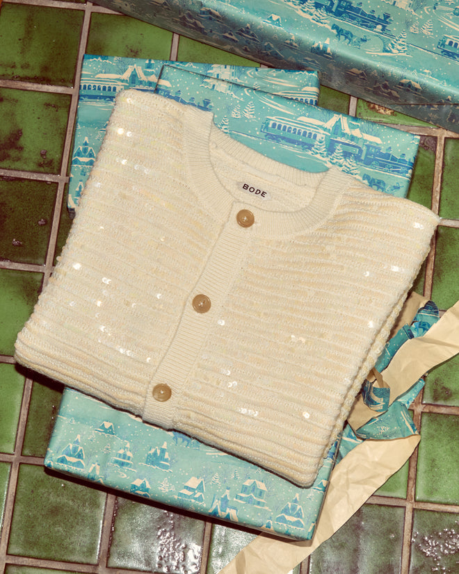 The Sequin Spotlight Cardigan in Ivory is displayed atop blue gift-wrapped presents on a green tiled surface.