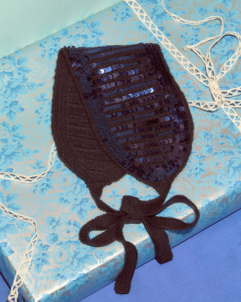The Sequin Spotlight Headband, a black sequined knit piece with a ribbon accent, is displayed on blue floral-patterned wrapping paper—a chic upgrade to your hair accessories collection.