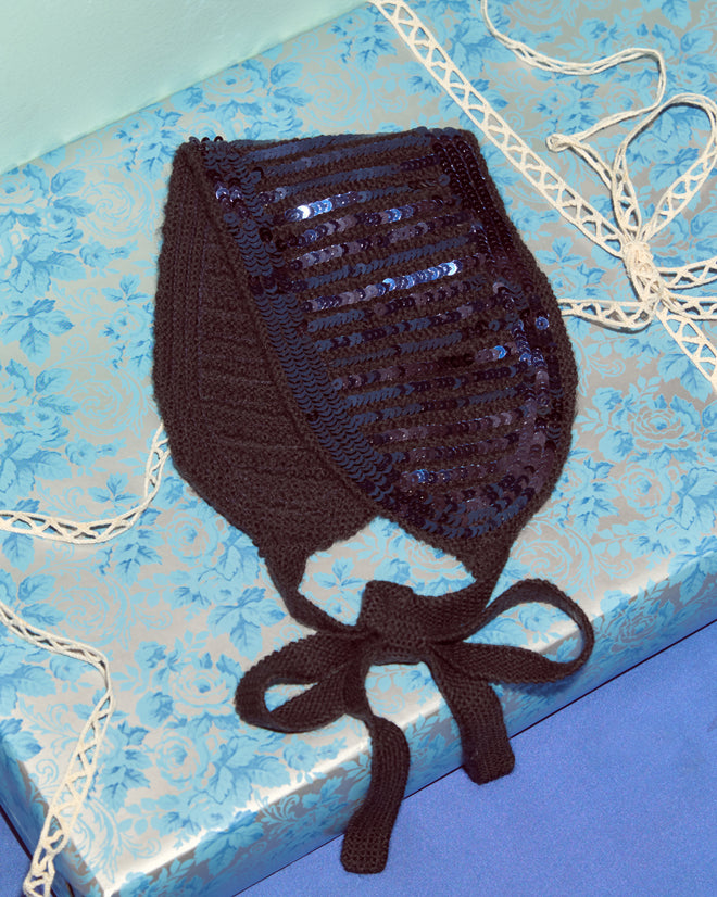 The Sequin Spotlight Headband, a black sequined knit piece with a ribbon accent, is displayed on blue floral-patterned wrapping paper—a chic upgrade to your hair accessories collection.