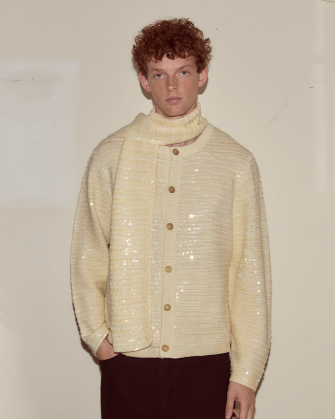 A person with curly hair wears a cream sequin cardigan and the Sequin Spotlight Scarf - Ivory, crafted from virgin wool, standing against a plain wall.
