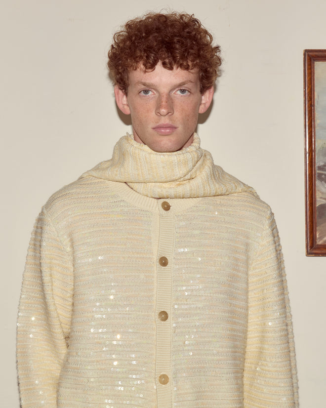 A young person with curly red hair stands indoors, elegantly wearing the Sequin Spotlight Scarf in ivory, paired with a cream sweater and handmade accessories.