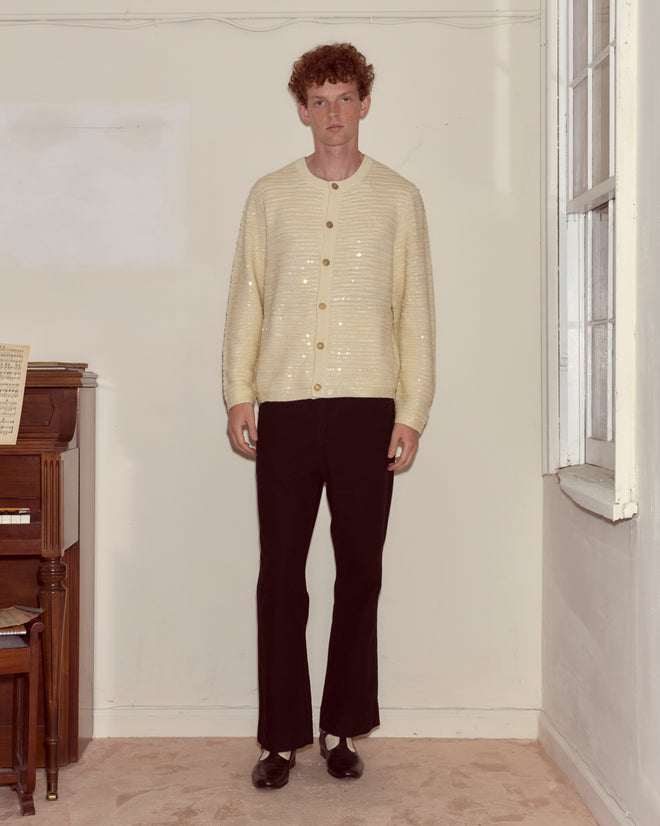 A person with curly hair stands indoors by a piano, wearing the Sequin Spotlight Cardigan in Ivory and black pants.