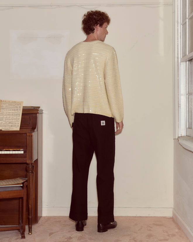 A person in an ivory Sequin Spotlight Cardigan and black pants stands with their back to the camera near a piano.