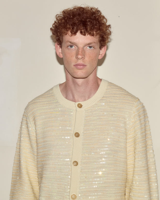 A young person with curly red hair wears the Sequin Spotlight Cardigan in Ivory, standing against a plain background.