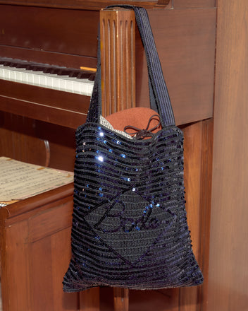 A Sequin Spotlight Tote - Black hangs on a piano, with papers and a brown folder visible inside.