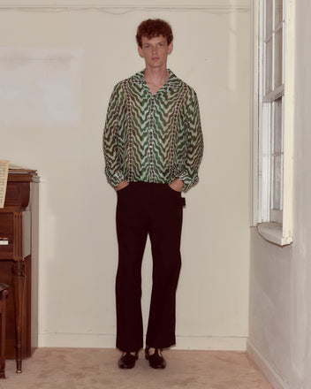 A person stands indoors by a window and piano, hands in pockets, wearing the Serpentine Long Sleeve—a green silk crepe shirt with a 1930s pattern—and black pants.