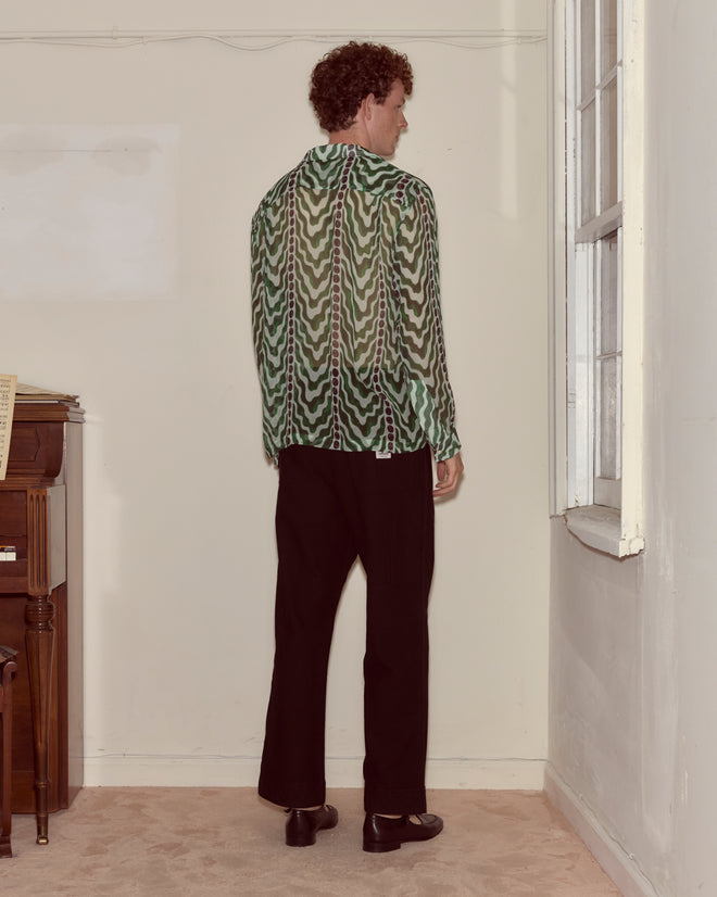 A person with curly hair stands indoors, facing a wall, wearing the green Serpentine Long Sleeve shirt and black pants.