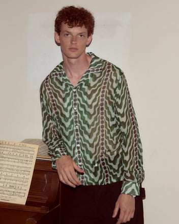 A man wearing the Serpentine Long Sleeve silk crepe shirt with a 1930s-inspired green pattern leans on a piano topped with sheet music, set against a plain wall.