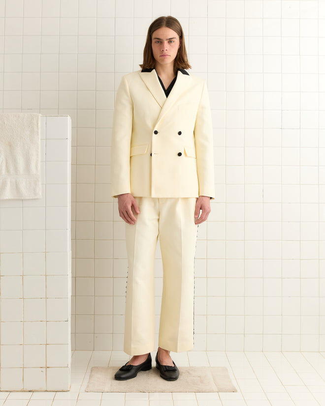 A person in a heavyweight cream Seymour Double-Breasted Suit Jacket and black shoes stands in a tiled room.