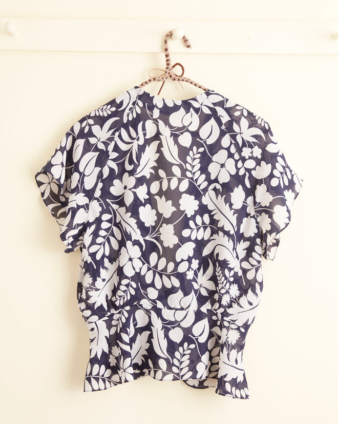 The Shadowed Ferns Top - 2, a navy and white floral blouse made from deadstock dress fabric, hangs on a hook against a cream-colored wall.