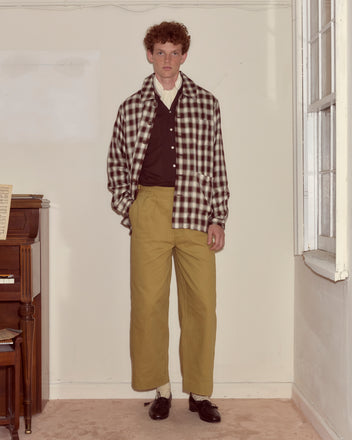 A man stands indoors by a piano, wearing the Shadow Plaid Franck Jacket with a brown shirt, tan pants, and black shoes.