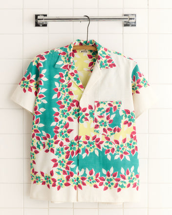 The Shamrock Bloom Shirt - S/M, featuring a vibrant green and red floral print, short sleeves, and vintage-inspired style, hangs on a metal hanger against a tiled white wall—a unique statement piece.