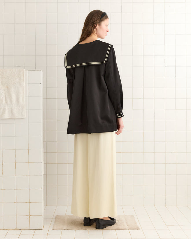 A woman wearing the Sheen Sailor Coat, featuring an ivory-trimmed black sailor-style top and paired with cream wide-leg pants, stands in a tiled room facing the wall.