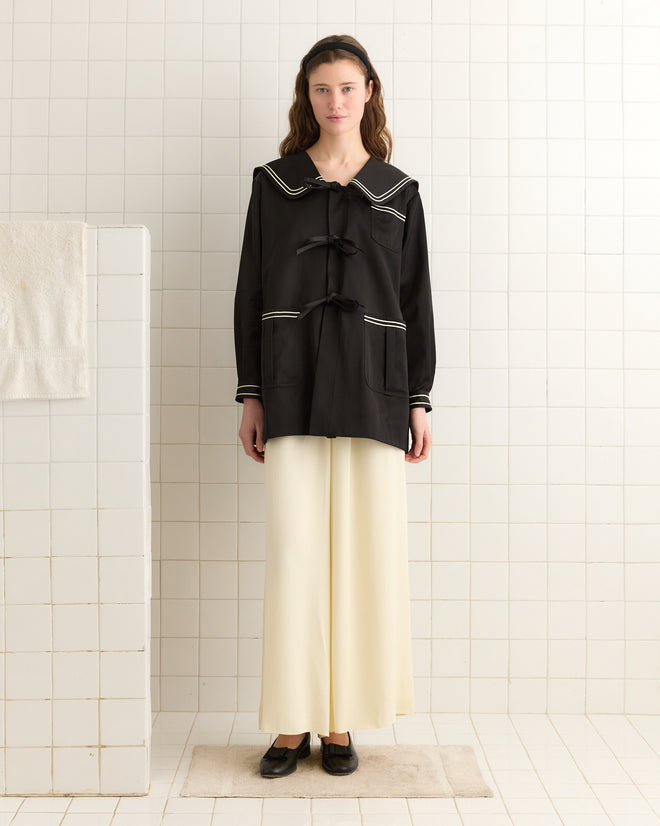 A person in a Sheen Sailor Coat and cream pants stands in a tiled room, with a towel hanging nearby.