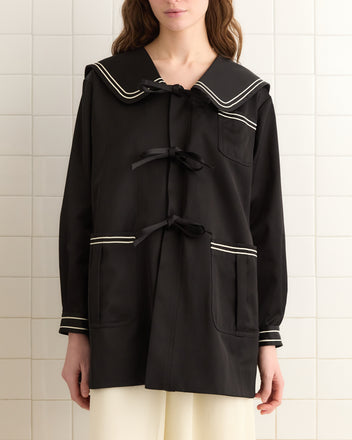 A person stands against a tiled wall wearing the Sheen Sailor Coat, a chic black outerwear piece with elegant white trim and distinctive bow closures, exuding timeless style.