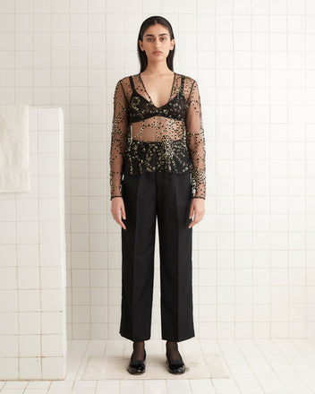 A person wearing the Sheer Fête Top, a sheer black and gold piece accented with gold foil appliqués, stands in a tiled room, paired with sleek black pants.