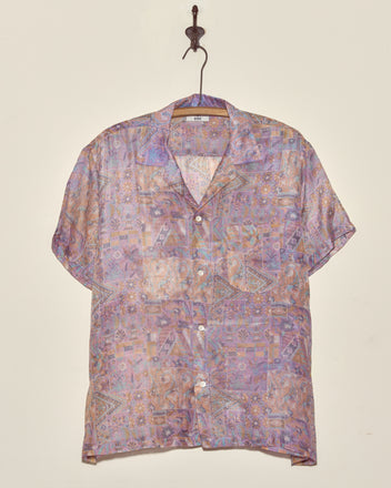 The Sheer Puzzle Shirt (M/L), a short-sleeved vintage top with a unique pattern in pastel shades, hangs on a wooden hanger against a plain wall.