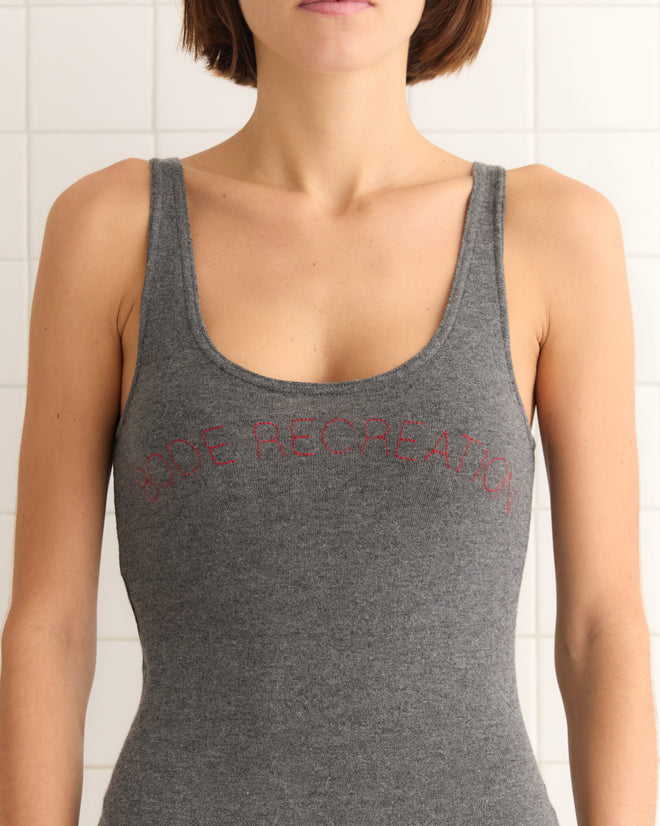 In a gray Shelly Romper, adorned with red "DOE RECREATION" text, someone stands against a white tiled wall, radiating casual style with an organic cotton vibe.