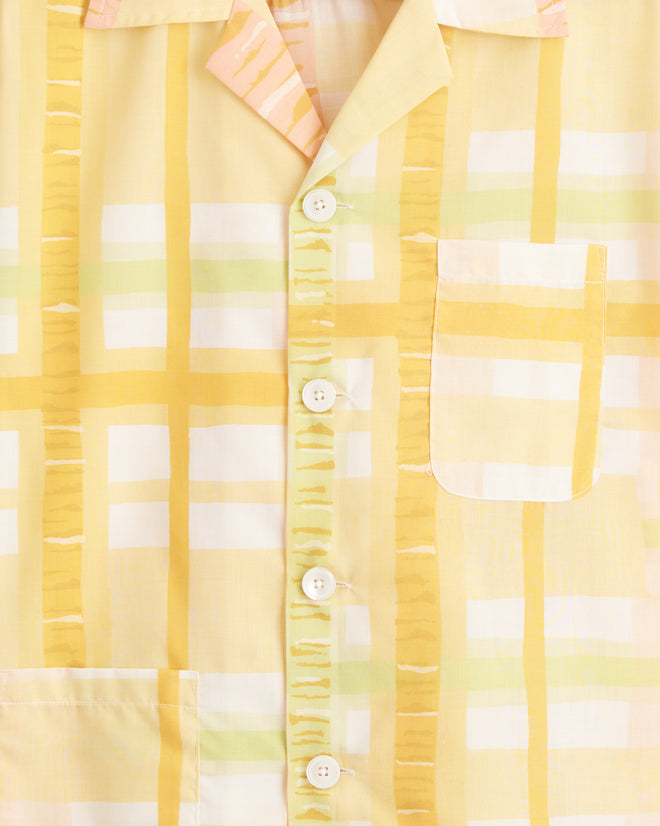 The Sherbet Plaid Shirt - XS/S, a unique short-sleeved button-up in yellow, white, and pink plaid, hangs on a hook against a cream-colored wall. This piece is crafted from deadstock yardage.