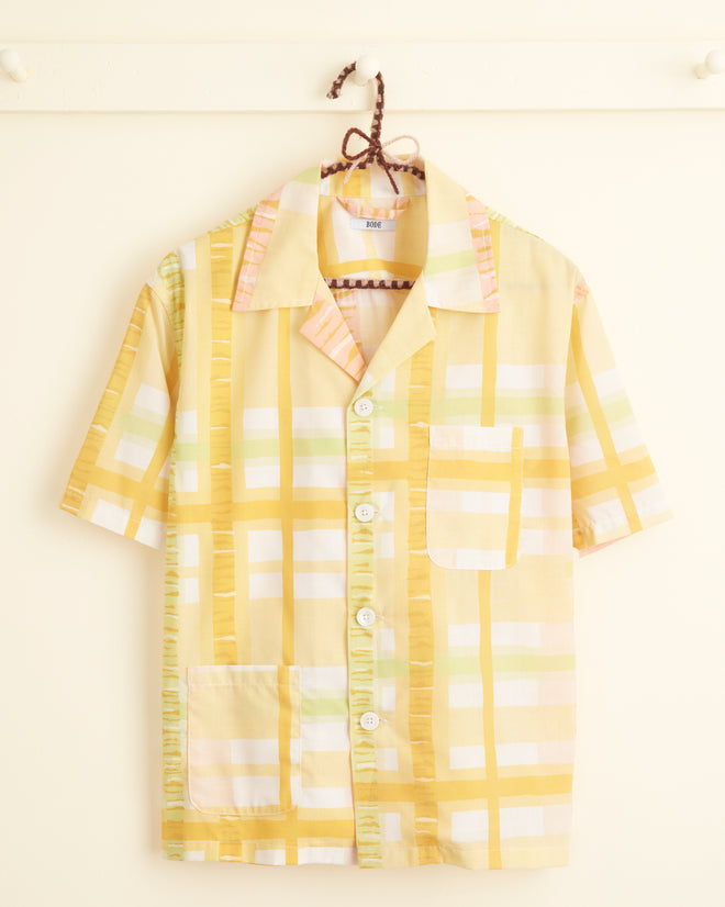 The Sherbet Plaid Shirt - XS/S, a unique short-sleeved button-up in yellow, white, and pink plaid, hangs on a hook against a cream-colored wall. This piece is crafted from deadstock yardage.