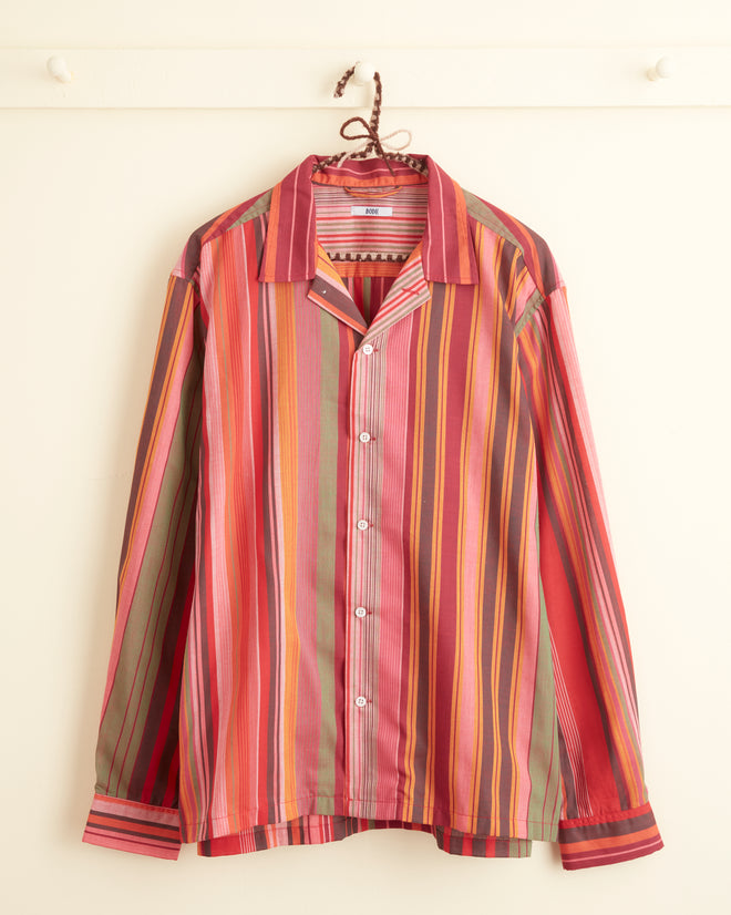 The Sherbet Stripe Shirt - XL/XXL, featuring a unique long-sleeved design with a vibrant stripe pattern, hangs on a hook against a light-colored wall.