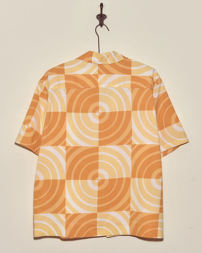 Sherbert Swirl Shirt - XXL: Short-sleeve vintage shirt featuring orange and yellow geometric circular patterns, displayed on a hook against a plain wall.