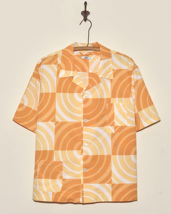 The Sherbert Swirl Shirt is a short-sleeve vintage top in orange and white, featuring a geometric circular pattern and displayed on a wall hook.