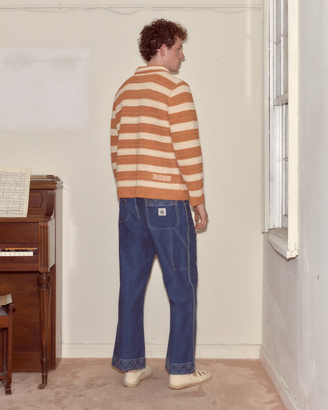 A person in a Sherbet Crochet Long Sleeve Shirt and jeans stands facing away in a room with a piano and a window.