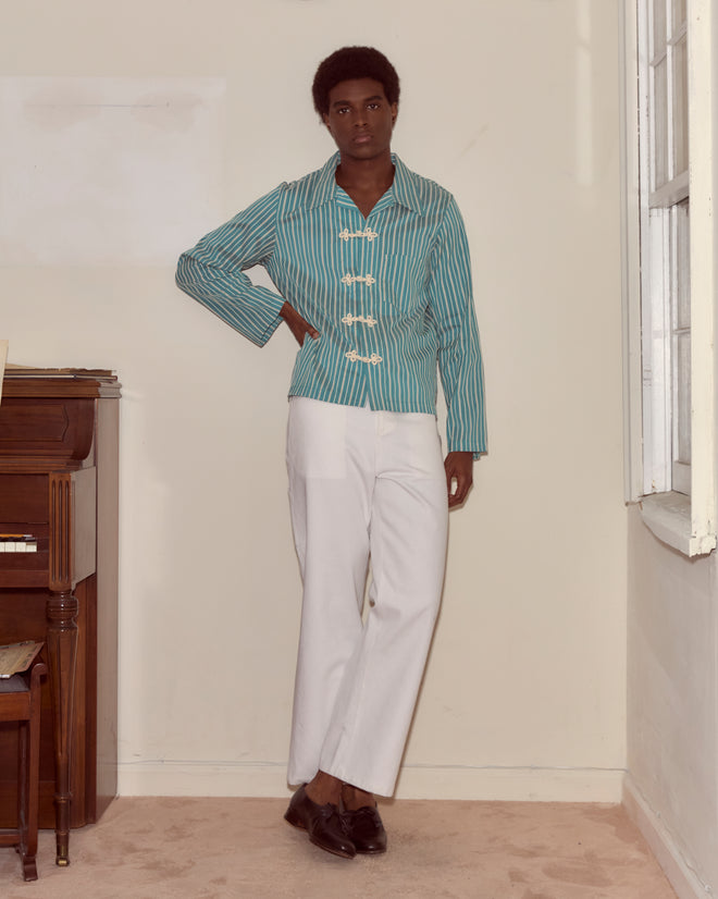 Indoors by a piano, a person wears the Shore Stripe Long Sleeve Shirt—a teal top with blue and white stripes inspired by 1940s men's styles—and white pants, looking at the camera.
