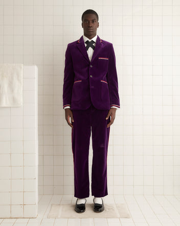 A man wearing a Showcase Suit Jacket in purple velvet with elegant soutache trim stands in a white tiled room next to a towel holder.