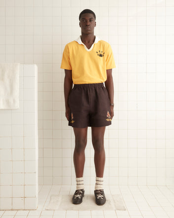 A man stands in a tiled room wearing a yellow shirt with a logo, 100% linen embroidered Show Pony Shorts, patterned socks, and black shoes.