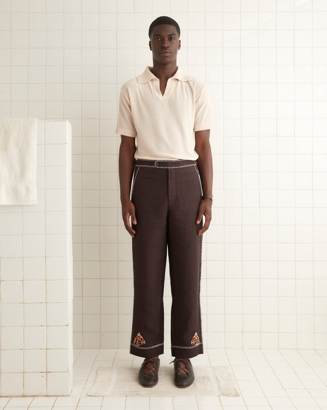 A person stands against a tiled wall wearing a light-colored short-sleeve shirt and dark Show Pony Trousers adorned with small patches near the hem. They are wearing brown shoes and posed with arms to the side.