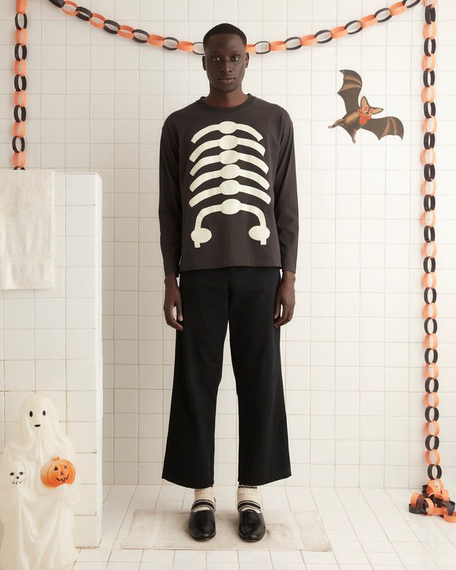 A Skeleton Long Sleeve Tee with a glow-in-the-dark skeleton print hangs on a wooden hanger, complemented by a vintage-style pumpkin face decoration on the wall behind it.