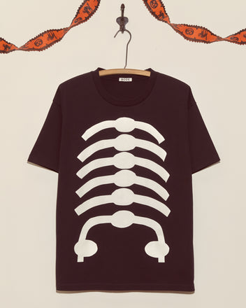 The Skeleton Tee, a black shirt with a white ribcage design, hangs on the wall with orange Halloween garland above—a vintage-style top ideal for fans of unique Halloween shirts.