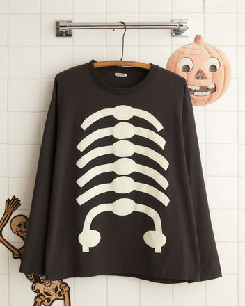 A Skeleton Long Sleeve Tee with a glow-in-the-dark skeleton print hangs on a wooden hanger, complemented by a vintage-style pumpkin face decoration on the wall behind it.