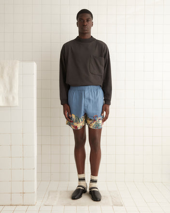 A person stands in a tiled room wearing a black long-sleeve shirt and Ski Lift Shorts with a tropical pattern. They have striped socks and black shoes on and stand on a white mat.