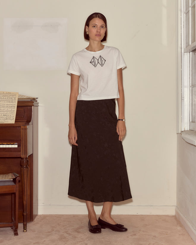 A woman stands indoors by a piano, wearing a white t-shirt and the Moonflower Jacquard Skirt in black, looking at the camera.