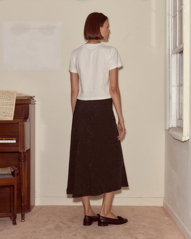 A woman stands indoors facing away, wearing a white top, the Moonflower Jacquard Skirt - Black, and brown shoes near a piano.