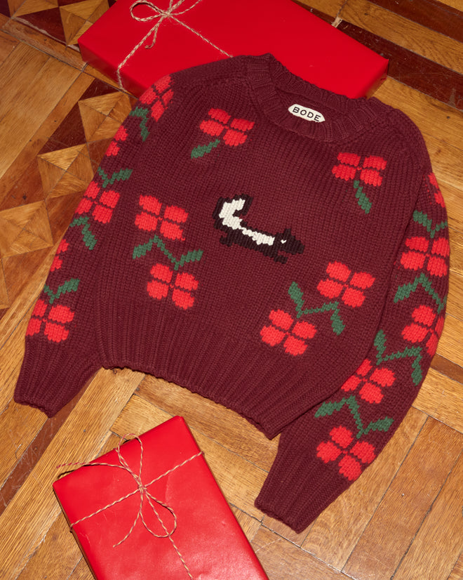 The Skunk Sweater is a maroon hand-knit sweater featuring red flowers and a black-and-white skunk, shown beside two wrapped red gifts on a wooden floor.