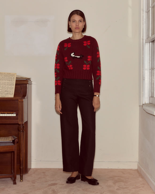 A woman wearing the Skunk Sweater, a chunky red wool sweater with a flower pattern, stands by a piano with sheet music on top, next to a window.