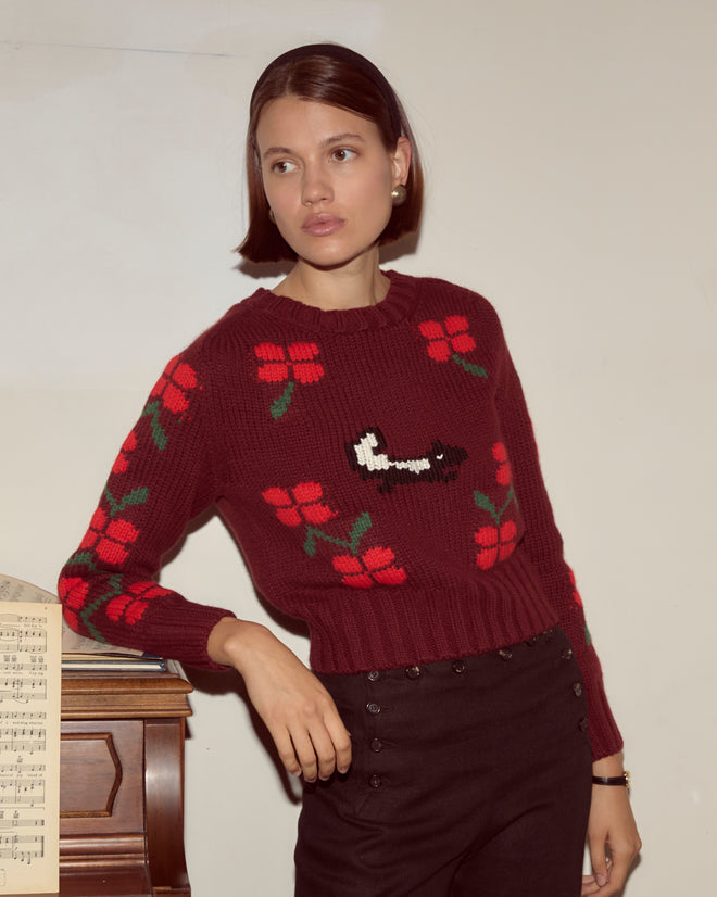 A woman in a Skunk Sweater, colored maroon with hand-knit floral details, leans against a piano with sheet music, gazing to the side.