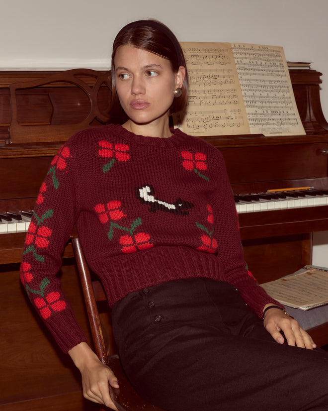 A woman wearing the Skunk Sweater, a hand-knit maroon piece with red flowers, sits in front of a piano with sheet music on the stand.