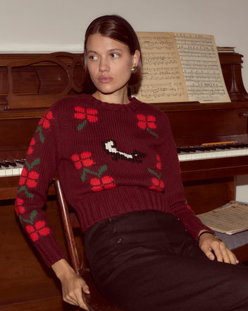 A woman wearing the Skunk Sweater, a hand-knit maroon piece with red flowers, sits in front of a piano with sheet music on the stand.