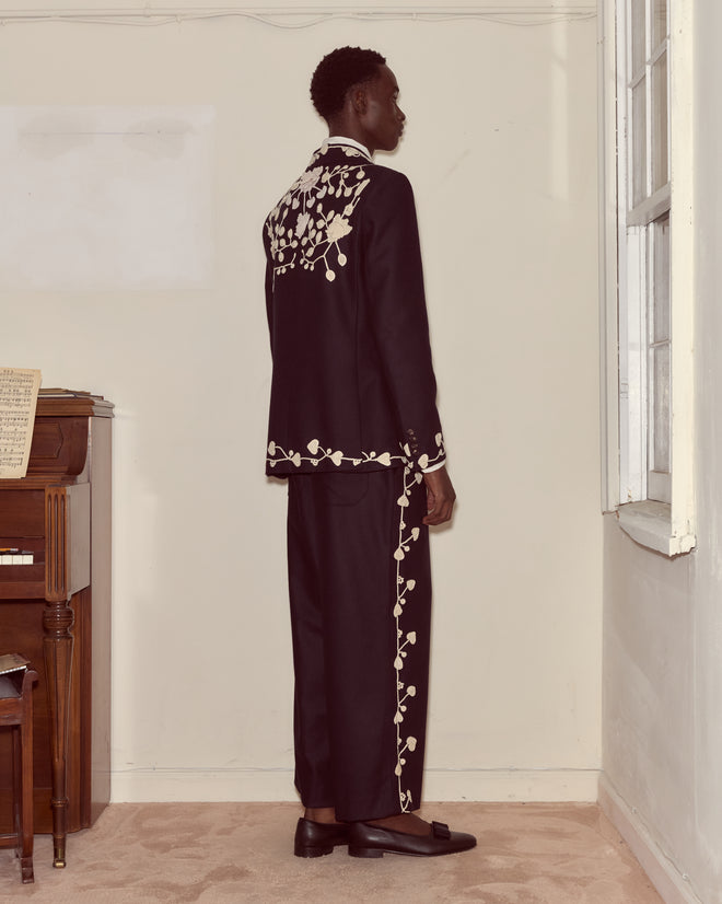 A person in a Snowdrop Appliqué Suit Jacket - Navy with white hand-crocheted floral embroidery stands sideways near a piano and a window.