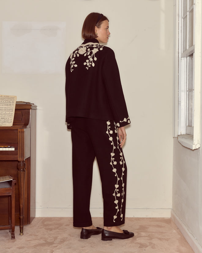A woman in the Snowdrop Appliqué Jacket stands near a piano, facing sideways by a window.